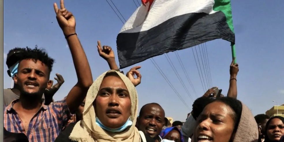 News Article A group of Sudanese people are gathered in protest, shouting. Some wear masks below their chin. In the back right, someone waves a Sudanese flag.