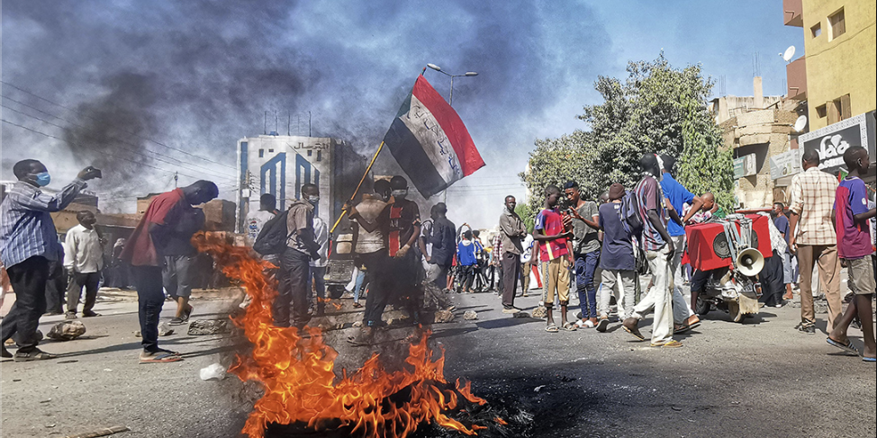 Screenshot 2025-03-29 at 10.50.52 AM A street fire toward the front of the frame is surrounded by people walking in the street. Behind it, a young man in a mask and a football jersey waves a Sudanese flag