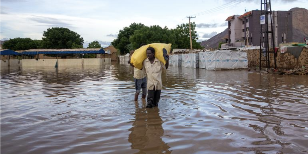 News Article A pair of young men walk through knee high water. The one in the front has a yellow sack perched on his shoulders and his sandals in his hand.