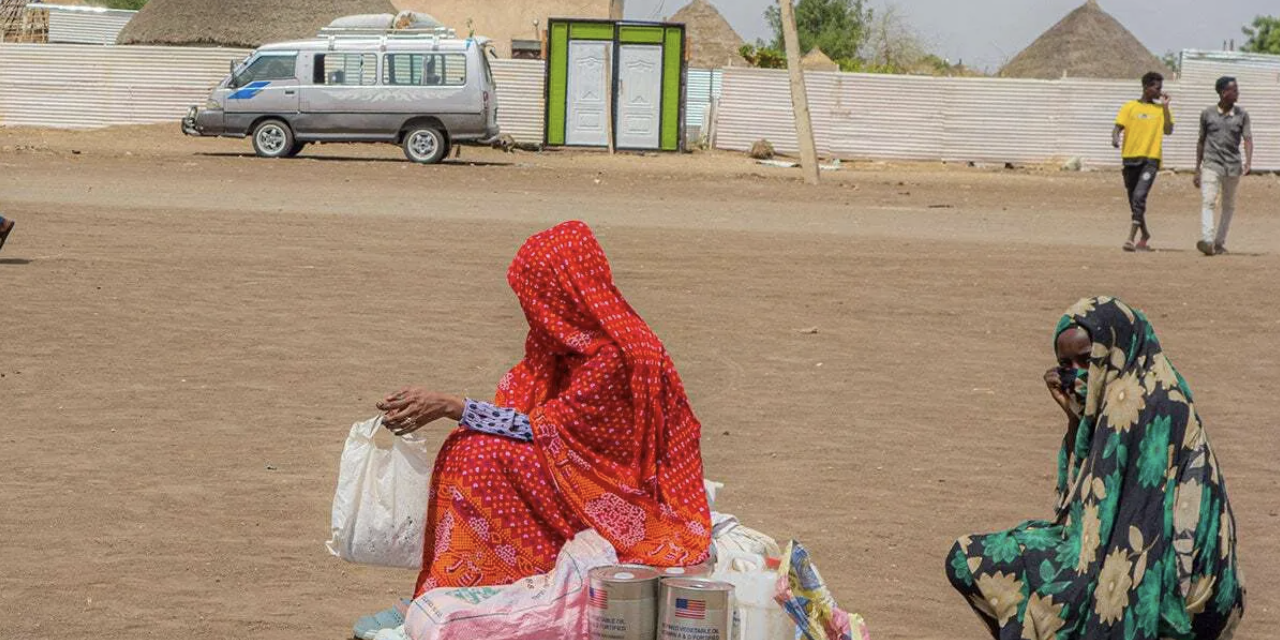 News and Stories Woman in burqas sit on their packages in an open sandy area, with a van visible in the background.
