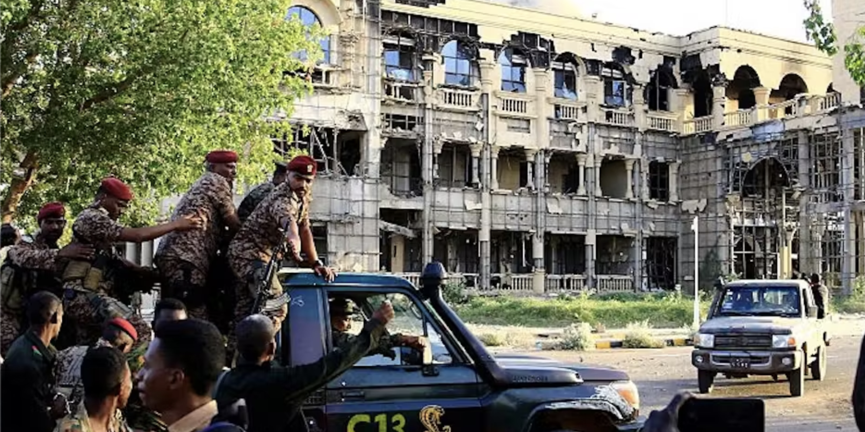 News and Stories Armed forces ride in the back of a pickup truck to the far left of the image. They're riding toward a building that has sustained some damage and is lined with scaffolding