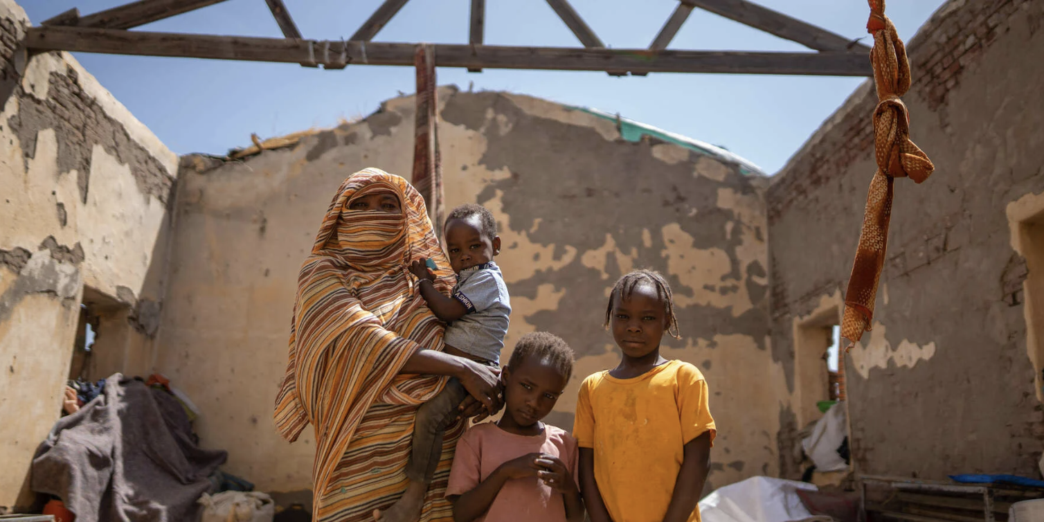 Screenshot 2025-03-29 at 2.43.38 PM A family with a woman in a burqa and three children. They stand under the frame of an open air building, with a roof seemingly being built above them.