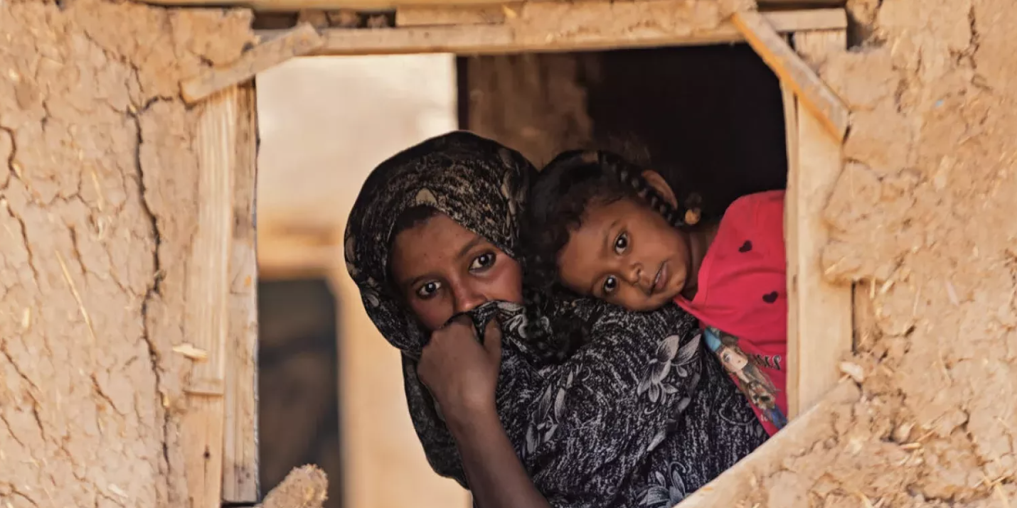 News and Stories A woman, wearing a hijab and carrying a young girl with braids and a red shirt on her back. The pair looks out from a window in a tan hut.