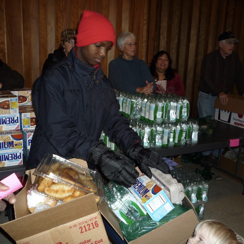 A group of people work the "line" of a food bank, handing out boxes of saltine crackers, bottles of water, eggs, and croissants.