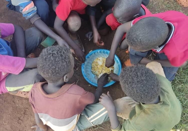 WhatsApp Image 2025-03-29 at 10.45.08 AM (3) Outside on a patch of grass and dirt, a group of young children gather around a bowl of food, sharing excitedly