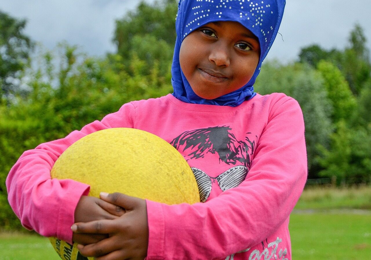 girl-4269839_1280 A young girl wearing hijab stands toward a camera, smiling slightly as she holds a yellow ball in her arms.