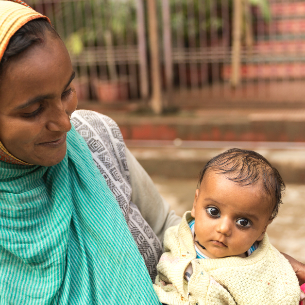 In front of a set of low steps, a woman wearing hijab looks attentively at a wide-eyed baby