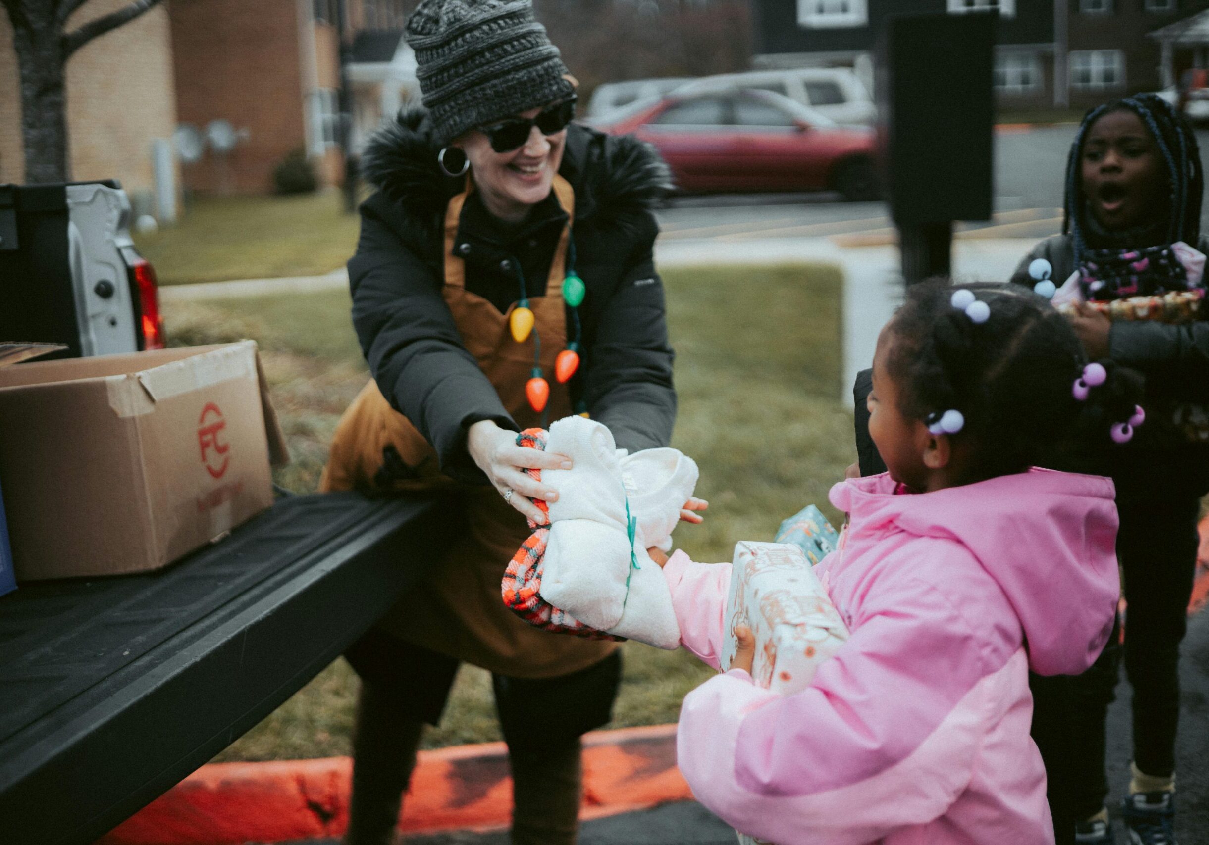 pexels-newmanphotographs-14831743 A young woman wearing a Christmas tree light necklace hands a package to a pair of young children